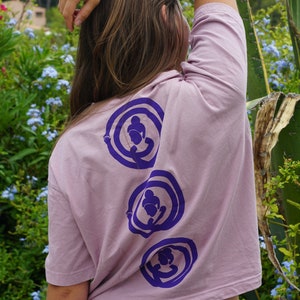 May include: A woman wearing a light pink, short-sleeved t-shirt with three purple circles on the back. Each circle has a stylized figure inside. The woman is wearing blue denim shorts.