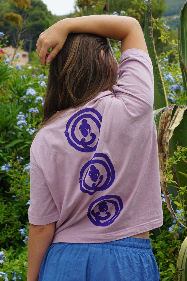 May include: A woman wearing a light pink, short-sleeved t-shirt with three purple circles on the back. Each circle has a stylized figure inside. The woman is wearing blue denim shorts.