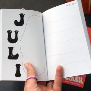 May include: An open notebook with the word "JULY" in black, stylized letters on the left page. The right page has horizontal lines. The word "Notizen" is at the top of the right page. A pink ring is visible.