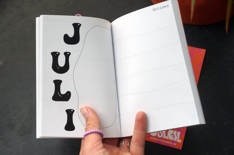 May include: An open notebook with the word "JULY" in black, stylized letters on the left page. The right page has horizontal lines. The word "Notizen" is at the top of the right page. A pink ring is visible.