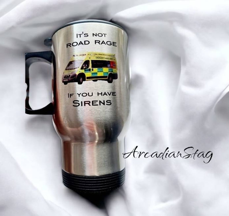 May include: A stainless steel travel mug with a black handle and lid. The mug has a graphic of an ambulance with the text "It's not road rage if you have sirens".