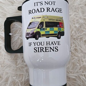 May include: White travel mug with a black lid and handle. The mug has a graphic of an ambulance with the text "It's not road rage if you have sirens".