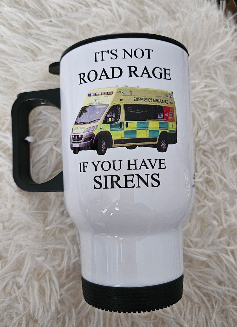 May include: White travel mug with a black lid and handle. The mug has a graphic of an ambulance with the text "It's not road rage if you have sirens".