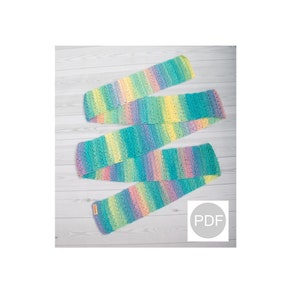 May include: A long, colorful crochet scarf with stripes of blue, green, yellow, pink, and purple. The scarf is folded and laid out on a white wooden surface. The text "PDF" is visible in a circle on the image.