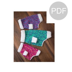 Könnte beinhalten: Drei gehäkelte Hunde-Pullover in Pink, Türkis und Lila mit weißen Rändern. Die Pullover liegen auf einer Holzoberfläche.