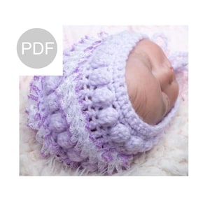 Puede incluir: Un gorro de bebé de ganchillo morado y blanco con ribete de encaje. El gorro tiene un lazo en la parte posterior.