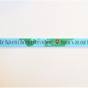 May include: A light blue wooden sign with a green and blue world map and a red pin. The sign reads "We haven't been everywhere, but it's on our list."