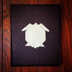 May include: A white origami turtle is displayed in a black frame against a dark wood background.