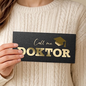 Könnte beinhalten: Eine schwarze Karte mit goldfarbenem Schriftzug "Call me DOKTOR" und einer goldenen Abschlusskappe. Die Karte wird von einer Person gehalten, die einen cremefarbenen Pullover trägt.