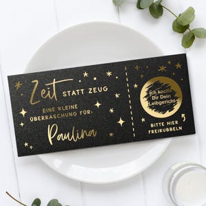 May include: A black gift certificate with gold lettering and star accents. The text includes "Zeit statt Zeug" and "Paulina". A scratch-off circle reveals "Ich koche Dir Dein Leibgericht". The certificate is on a white plate.