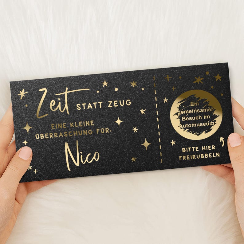 May include: A black gift card with gold foil text and star accents. The card reads "Zeit statt Zeug" and "Eine kleine &Uuml;berraschung f&uuml;r: Nico." A scratch-off area says "Ein gemeinsamer Besuch im Automuseum." The card is held by two hands.