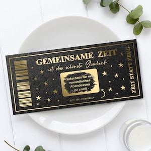 Könnte beinhalten: Ein schwarzer und goldener Gutschein mit dem Text "GEMEINSAME ZEIT ist das schönste Geschenk" und "ZEIT STATT ZEUG". Der Gutschein hat einen Rubbelbereich und Sternakzente. Er liegt auf einem weißen Teller.