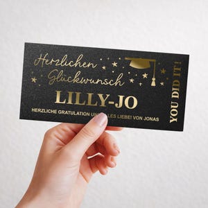 Könnte beinhalten: Eine schwarze Abschlusskarte mit goldfarbenen Buchstaben. Auf der Karte steht "Herzlichen Glückwunsch LILLY-JO" und "YOU DID IT!". Eine Grafik einer Abschlusskappe ist ebenfalls abgebildet. Die Karte wird von einer Person gehalten.