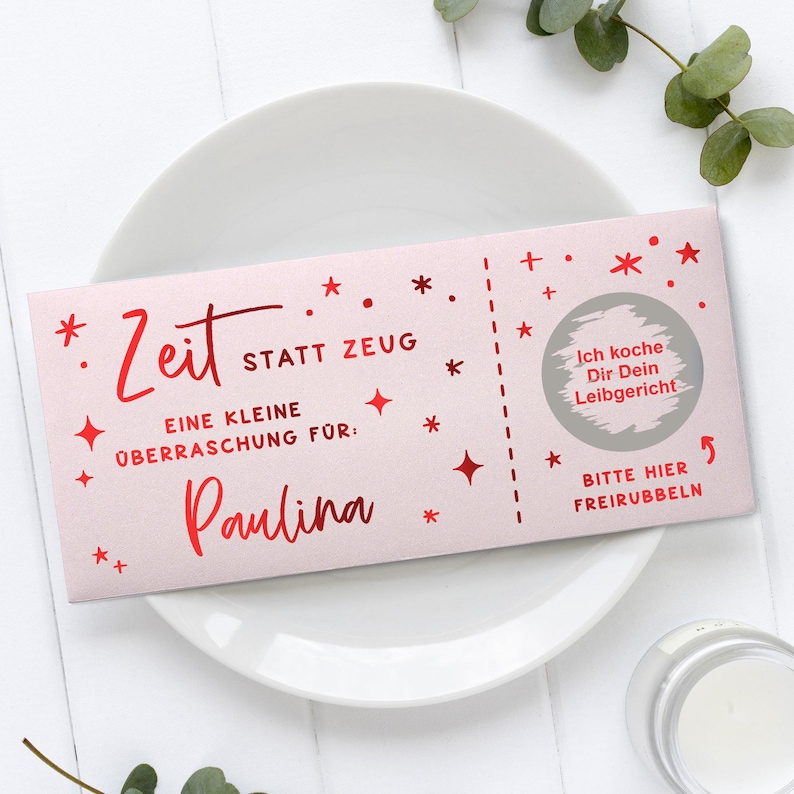 May include: A pink gift certificate with red text and star and plus symbols. The text reads "Zeit statt Zeug" and "Paulina" is written in cursive. A scratch-off area says "Ich koche Dir Dein Leibgericht".