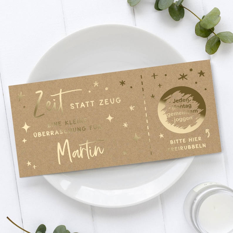 May include: A brown gift certificate with gold foil text and star accents. The text includes "Zeit statt Zeug" and "Martin". A scratch-off circle reads "Jeden Montag gemeinsam joggen". The certificate is on a white plate.