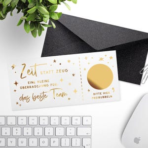 May include: A white card with gold foil text and a scratch-off circle, accompanied by a black envelope. The card reads "Zeit statt Zeug" and "das beste Team". A keyboard and mouse are in the background.