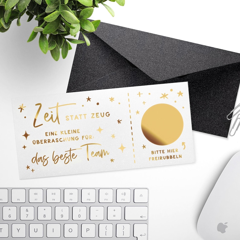 May include: A white card with gold foil text and a scratch-off circle, accompanied by a black envelope. The card reads "Zeit statt Zeug" and "das beste Team". A keyboard and mouse are in the background.