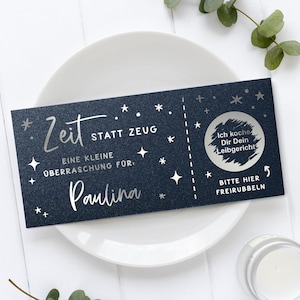 May include: A dark blue gift certificate with silver text and star accents, resting on a white plate. The text includes "Zeit statt Zeug" and "Paulina". A scratch-off area says "Ich koche Dir Dein Leibgericht".