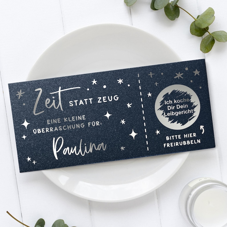 May include: A dark blue gift certificate with silver text and star accents, resting on a white plate. The text includes "Zeit statt Zeug" and "Paulina". A scratch-off area says "Ich koche Dir Dein Leibgericht".