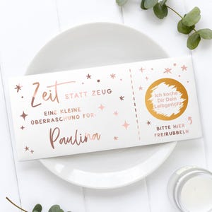 May include: A white gift certificate with rose gold text and star accents, resting on a white plate. The text includes "Zeit statt Zeug" and "Paulina". A gold circle with German text is on the right side.