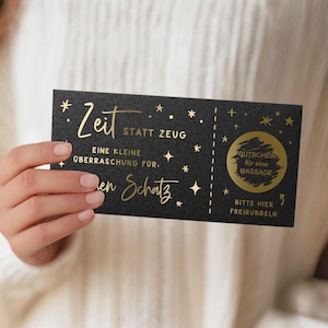 May include: A black gift certificate with gold foil text and star accents. The certificate reads "Zeit statt Zeug" and "Gutschein f&uuml;r eine Massage". The certificate is held by a person.