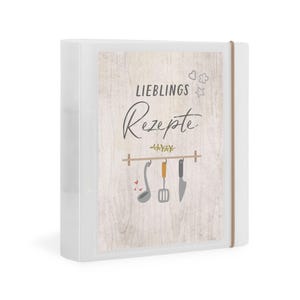 Könnte beinhalten: Ein weißer Ordner mit den Worten "LIEBLINGS Rezepte" auf der Vorderseite. Das Cover zeigt einen Holzgrund mit Illustrationen von Küchenutensilien. Ein braunes Gummiband sichert den Ordner.