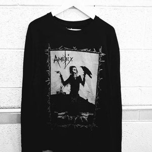 May include: Black long-sleeve shirt with a graphic print of a person holding a bird. The graphic is in a white rectangle with a black barbed wire border. The text "Amebix" is printed in white above the graphic.
