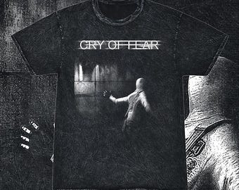 May include: A black short-sleeve t-shirt with a distressed texture. The front features the white text "CRY OF FEAR" above a graphic of a hooded figure in a dark, shadowed environment with a wall and light source, rendered in a grainy, high-contrast style.