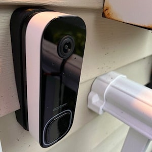 May include: A white and black doorbell camera with a black button and the word "ecobee" printed on it. The camera is mounted on a white wall.