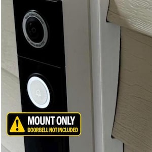 May include: A white angled mounting bracket for a video doorbell, featuring a rectangular shape that attaches to a wall. The bracket is shown with a black video doorbell attached, which displays the text MOUNT ONLY DOORBELL NOT INCLUDED.