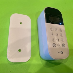 May include: A white wall mount with two holes and a white doorbell with a keypad and a blue light. The keypad has numbers 1 through 9, 0, a phone icon, and a house icon. The doorbell has the text "myQ" on the bottom.
