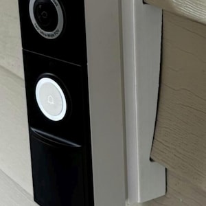 Puede incluir: Un timbre con video en blanco y negro montado en una pared de color claro. El timbre tiene una cámara, un botón y el texto "True Video Doorbell". El timbre está sujeto a un soporte de montaje blanco.