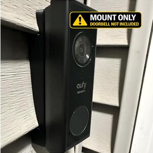 May include: A black plastic mounting bracket for a video doorbell, featuring a rectangular shape and a flat surface. A yellow warning label at the top reads MOUNT ONLY DOORBELL NOT INCLUDED. The mount is attached to a light-colored exterior wall.