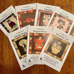 May include: A stack of eight colorful quilting patterns featuring cats and vegetables. The patterns are titled "Eggplant Purr-mesan", "Yammy Cat", "Mushkit", "Broc-Kitty", "Pickle PusS", "Pepper Puss", "Brussels Cat", and "Garden Patch Cats".