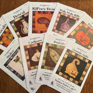May include: A collection of quilt pattern booklets featuring whimsical cat designs. Titles include "Avocato," "Boscat," "Kitt'ney Bear," and "Gourdo Gato." Each booklet showcases a colorful cat illustration within a patchwork border. The booklets are arranged on a wooden surface.