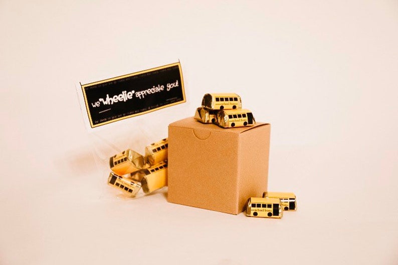May include: A small cardboard box filled with miniature yellow school bus chocolates. The box is topped with a stack of additional bus chocolates. A clear plastic bag with more bus chocolates and a black tag with the text "we "wheelie" appreciate you" is in the background.