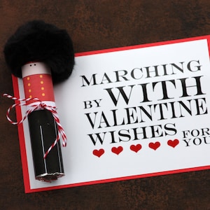 Op de afbeelding: Een Valentijnsdagkaart met een rode rand en de tekst "MARCHING BY WITH VALENTINE WISHES FOR YOU" staat naast een decoratieve soldaat. De soldaat is gemaakt van een zwarte buis, rood en wit papier en een zwarte, harige hoed.