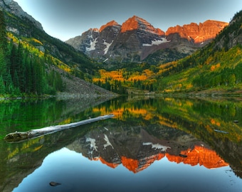 Puede incluir: Una vista panorámica de una cordillera con un lago en primer plano. Las montañas están cubiertas de árboles y nieve, y el lago refleja las montañas y el cielo. Un tronco caído se encuentra en el agua.