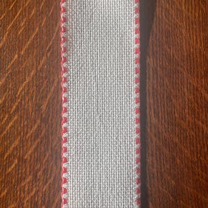 May include: A rectangular piece of white fabric with a textured weave. The edges are finished with a red, decorative stitch. The fabric is displayed on a wooden surface.