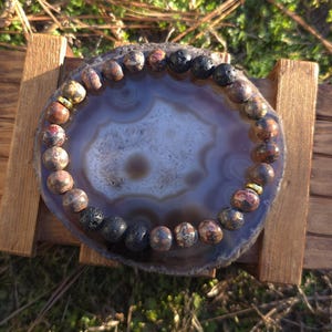 May include: A beaded bracelet with brown, black, and red stones rests on a polished agate slice. The agate is set on a small wooden display stand, with a natural outdoor background.
