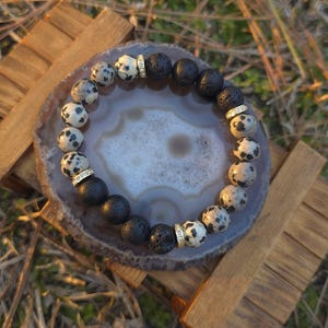 May include: A beaded bracelet featuring a mix of black lava and dalmatian jasper beads. The bracelet is accented with gold-colored spacers. The bracelet is displayed on a gray and white agate slice, resting on a wooden stand.