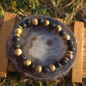 Op de afbeelding: Een kralenarmband met afwisselend zwarte lavasteen en bruinbeige zandsteen kralen, geaccentueerd met goudkleurige afstandhouders. De armband wordt gepresenteerd op een grijs-witte agaat schijf, rustend op een houten standaard, met een natuurlijke buitenachtergrond.