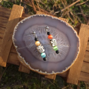May include: A pair of dangle earrings featuring silver owl charms and colorful beads. The earrings have a post-back closure and are displayed on a gray and white agate slice. The beads are in shades of orange, green, blue, yellow, and black.