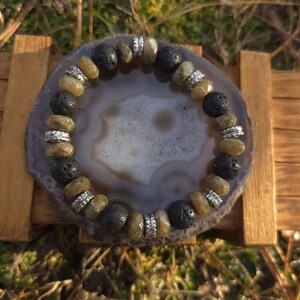 Puede incluir: Una pulsera de cuentas con cuentas de roca volcánica negra, cuentas de piedras preciosas de color marrón claro y cuentas espaciadoras plateadas. La pulsera se exhibe sobre una rodaja de ágata gris y blanca, apoyada en un pequeño soporte de madera.
