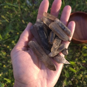 Puede incluir: Un puñado de cristales de cuarzo ahumado en varios tamaños y tonos de marrón y gris. Los cristales se sostienen en una mano abierta sobre un fondo de hierba verde y un cuenco de madera.