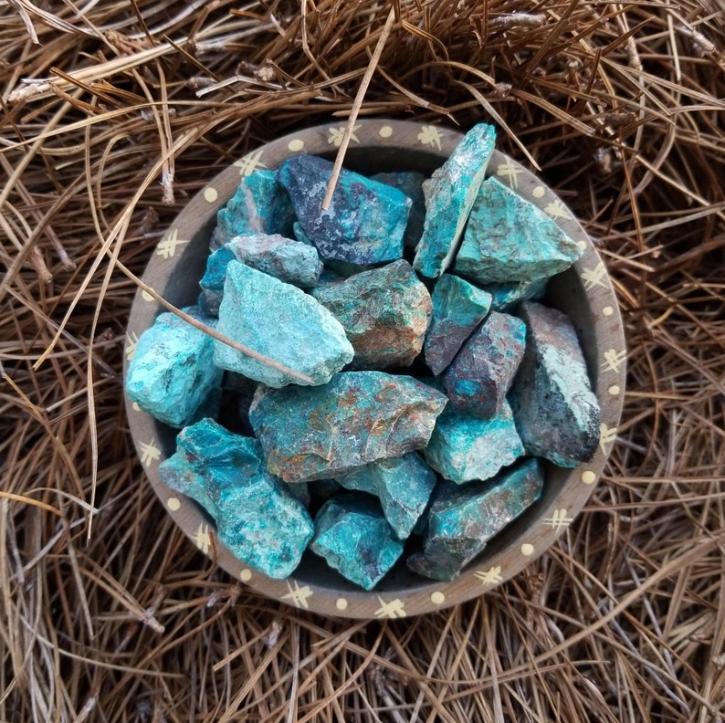 A tribal marked bowl sitting in a patch of pine needles full of rough chunks of vibrant blue stones.