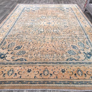 Op de afbeelding: Een groot, rechthoekig tapijt met een beige achtergrond en ingewikkelde blauwe bloemmotieven. Het tapijt heeft een rand met een herhalend patroon van blauw en beige.
