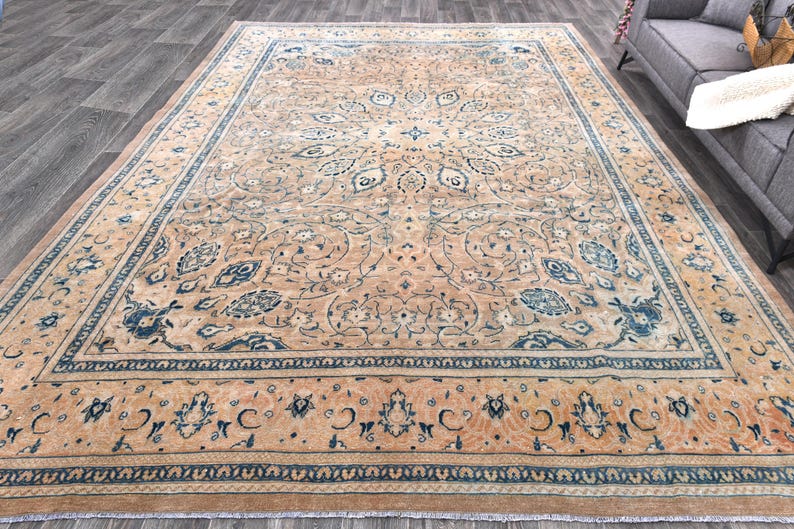 Op de afbeelding: Een groot, rechthoekig tapijt met een beige achtergrond en ingewikkelde blauwe bloemmotieven. Het tapijt heeft een rand met een herhalend patroon van blauw en beige.