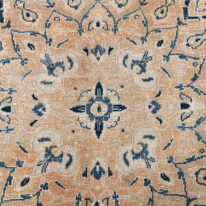 Op de afbeelding: Een close-up van een beige kleed met een blauw en wit geometrisch patroon. Het kleed heeft een vervaagde, vintage uitstraling.