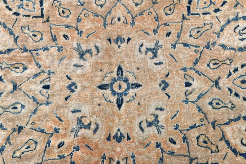Op de afbeelding: Een close-up van een beige kleed met een blauw en wit geometrisch patroon. Het kleed heeft een vervaagde, vintage uitstraling.